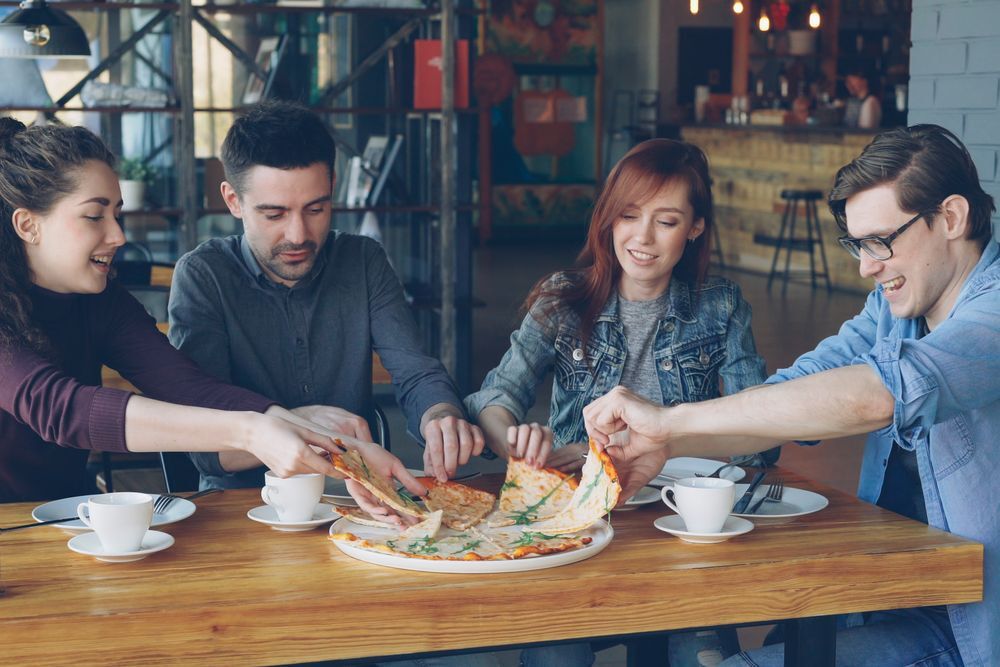 Front-facing image of four friends gathered around at a table in a restaurant in Montgomery Township, New Jersey, and sharing a pizza together. 